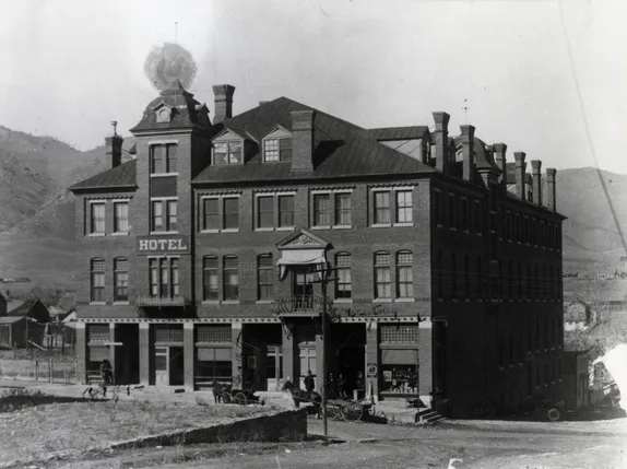 3 story brick building with sign saying "HOTEL" square turrets in front and back and many chimneys.  Mountains in background.