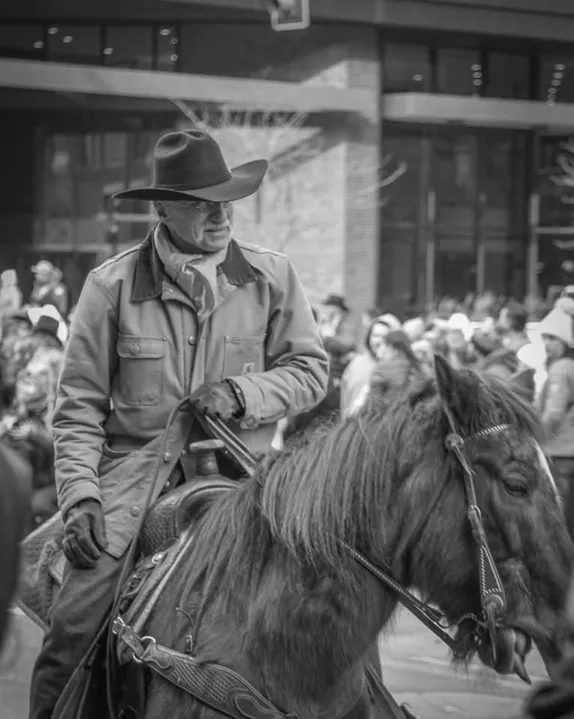 black & white image of a man in a cowboy hat & Carhartt jacket on a brown horse. Crowd along the curb in the background.