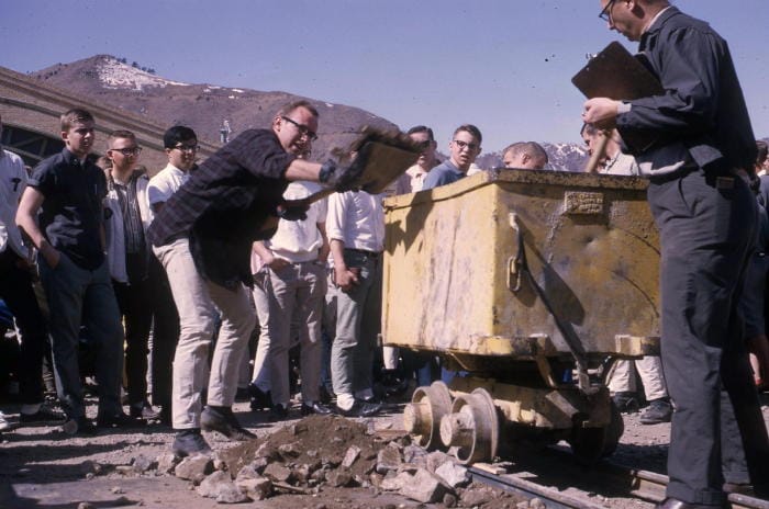 Young man shovels dirt and rocks into an ore cart while a crowd watches.