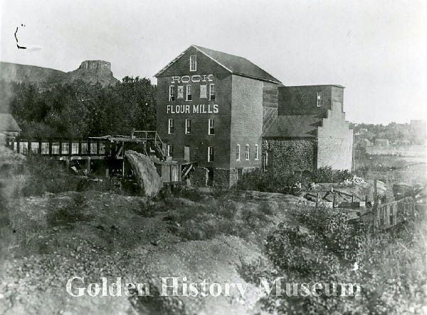 photo showing the Rock Flower Mill on 9th Street with water in the flume and Castle Rock in the background