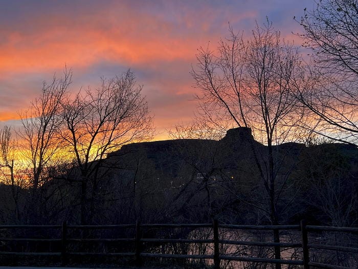 colorful sunrise silhouettes South Table Mountain and several bare trees. Clear Creek is faintly visible in the foreground.