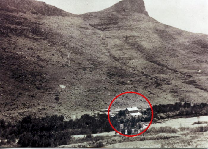 Black and white photo shows a small building surrounded by trees on the south bank of Clear Creek.  Castle Rock appears above the building