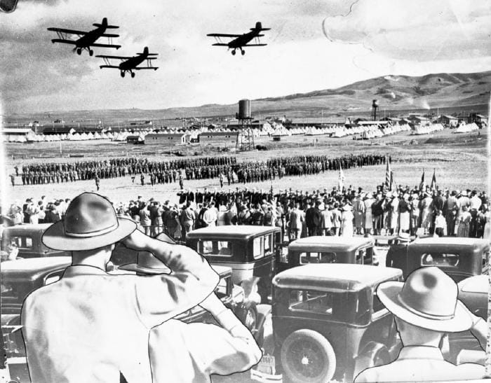 Military maneuvers at Camp George West in 1933.  Saluting soldiers in the foreground, troops in formation, canvas tents, and South Table Mountain in the background, and biplanes flying overhead.