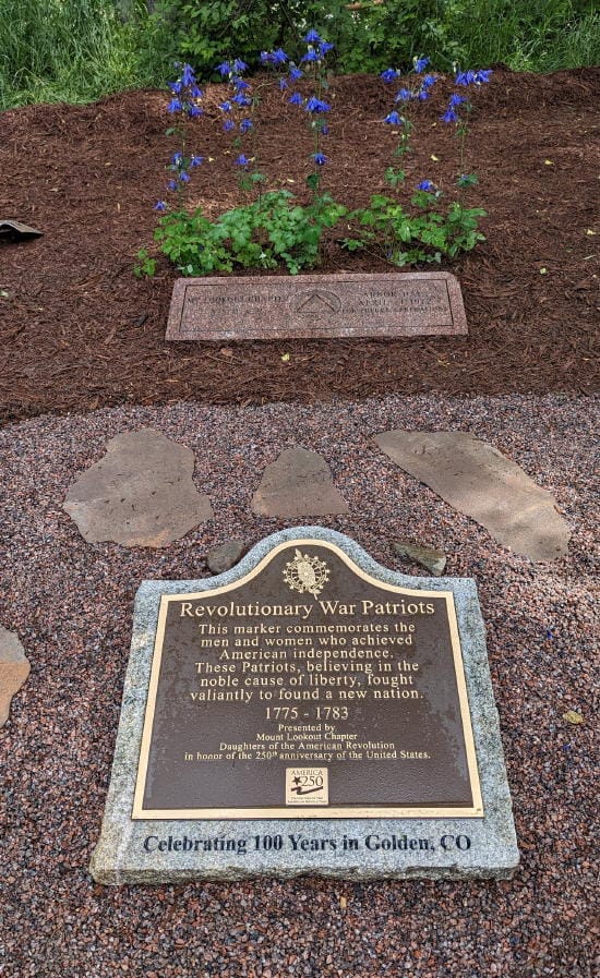 A brass plaque honoring Revolutionary War patriots, on a gray granite base.  Donated by the Lookout Mountain Chapter, Daughters of the American Revolution, in celebration of their 100th anniversary.  A brown granite monument is nearby, commemorating the group's work in planting trees and establishing a park in that area.  A group of blue columbines is planted nearby.