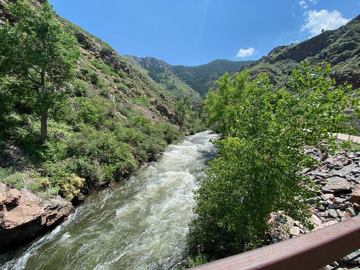 a view of Clear Creek Canyon taken from a bridge, looking down on the creek, which is very high. The foliage is all very green.