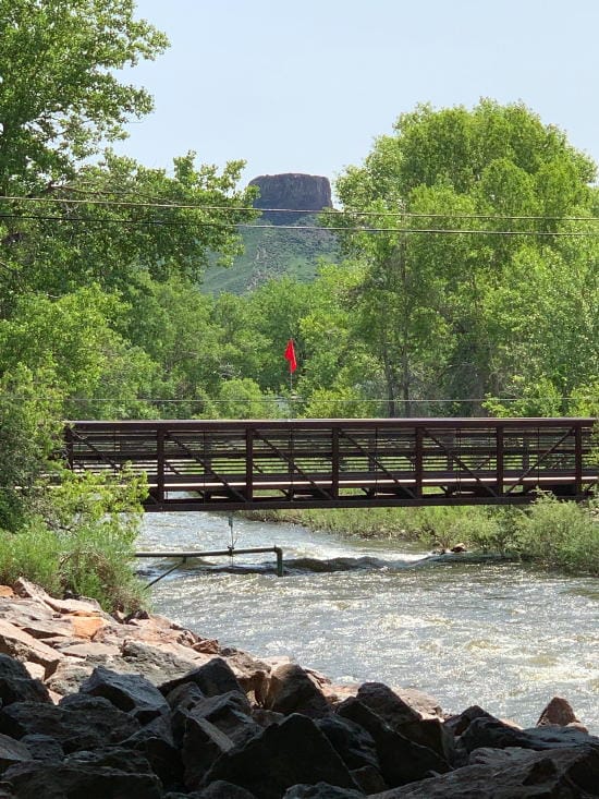 A red flag hangs flies above a bridge over Clear Creek, signally dangerously high, fast water.  Castle Rock in the background.