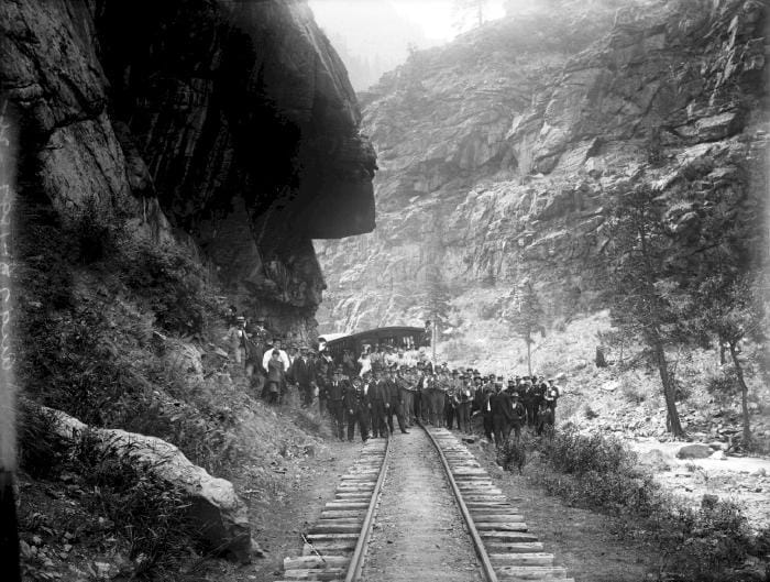 A group of well-dressed people pose in front of a seam locomotive in Clear Creek Canyon. A brass band, in uniform with instruments, in included in the crowd.