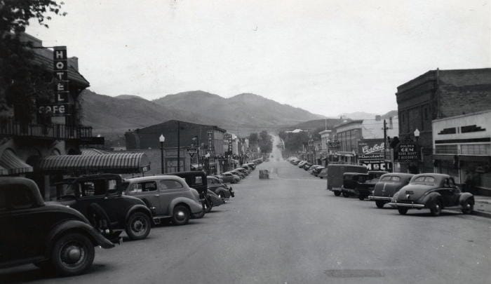 Black and white photo showing the commercial district of Washington Avenue in Golden Colorado.  Cars at diagonally parked on both sides of the street.  Many signs jut out from the buildings, including Hotel/Cafe, Conoco, Foss Drugs, Alpine Drugs, Golden Gem, Chocolate Shop, and Central Light and Power