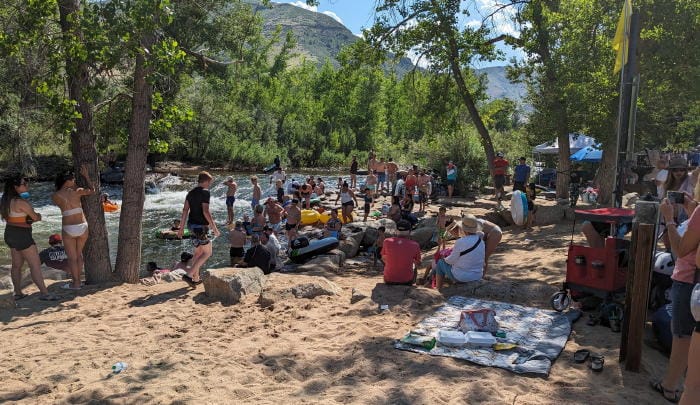 Large group of swimsuit-clad people on the "beach" section of Clear Creek in Golden.  Lots of rafts and tubes. Mt. Zion in the background.