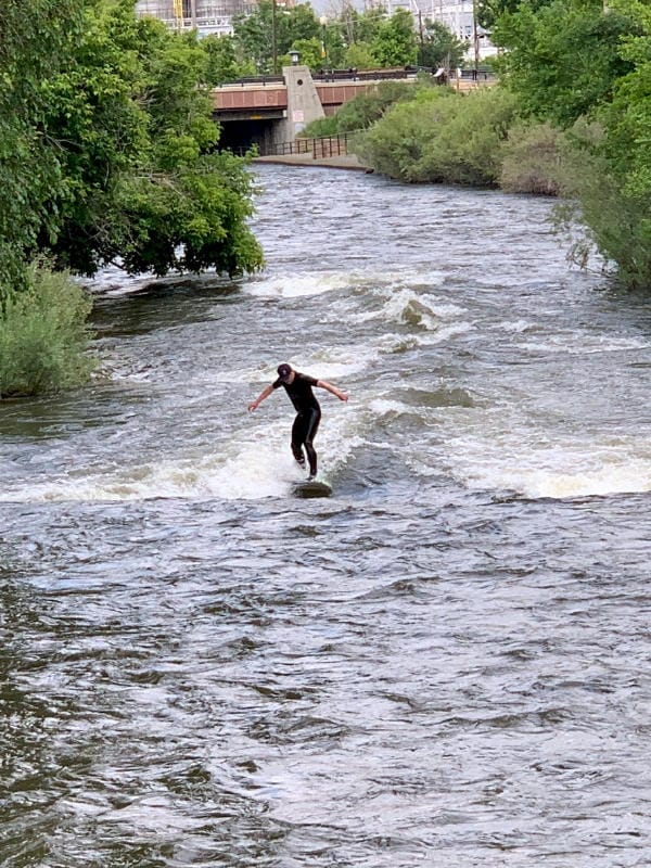 A person in a wet suit surfing on Clear Creek.  The water level is high.  The Ford Street appears in the background.