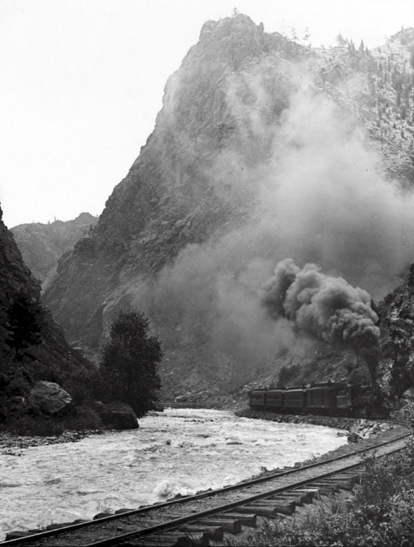 A coal-powered passenger train makes its way through Clear Creek canyon.  Clear Creek flows next to the tracks.