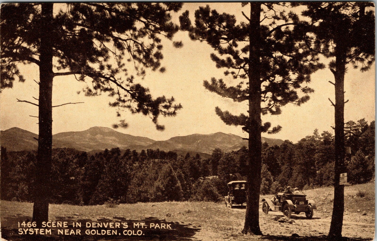 two 1920s-era cars on a narrow dirt road, heading into trees, with mountains in the background