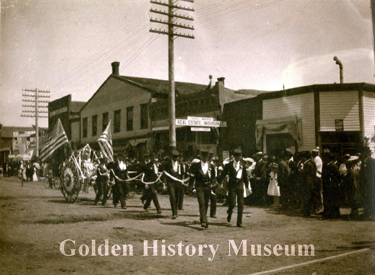 team of men in uniforms pulling a hose cart that is decorated with American flags - business buildings in background