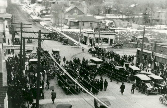 black and white photo showing a large crowd of people with cars and flags on Washington Avenue