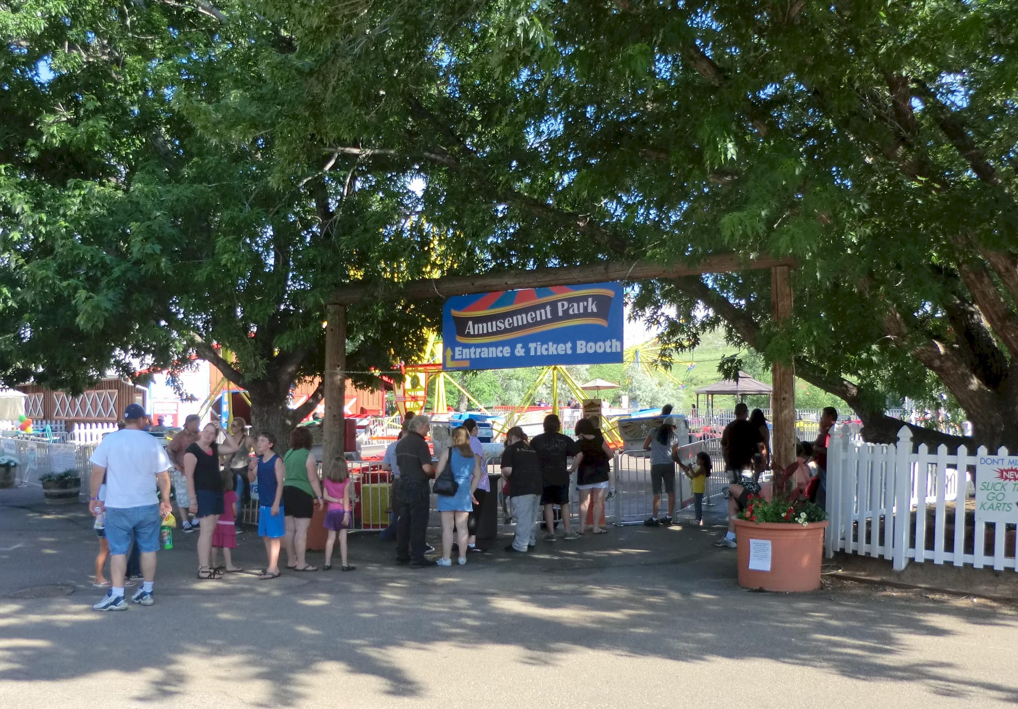 many people near an entrance gate to an area surrounded by a white picket fence - sign announces "Amusement Park"
