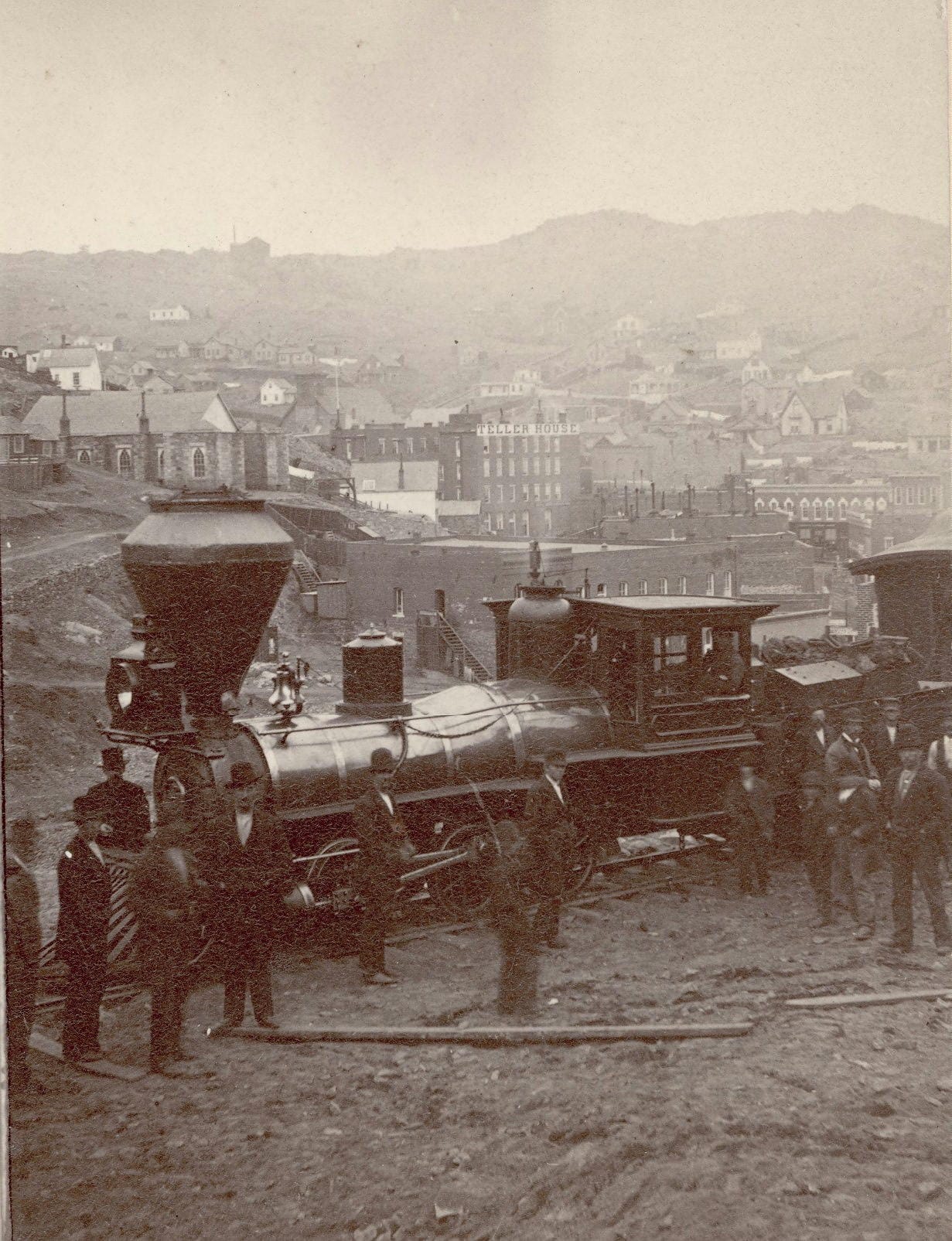 group of men in suits and hats standing around a locomotive with Central City in background