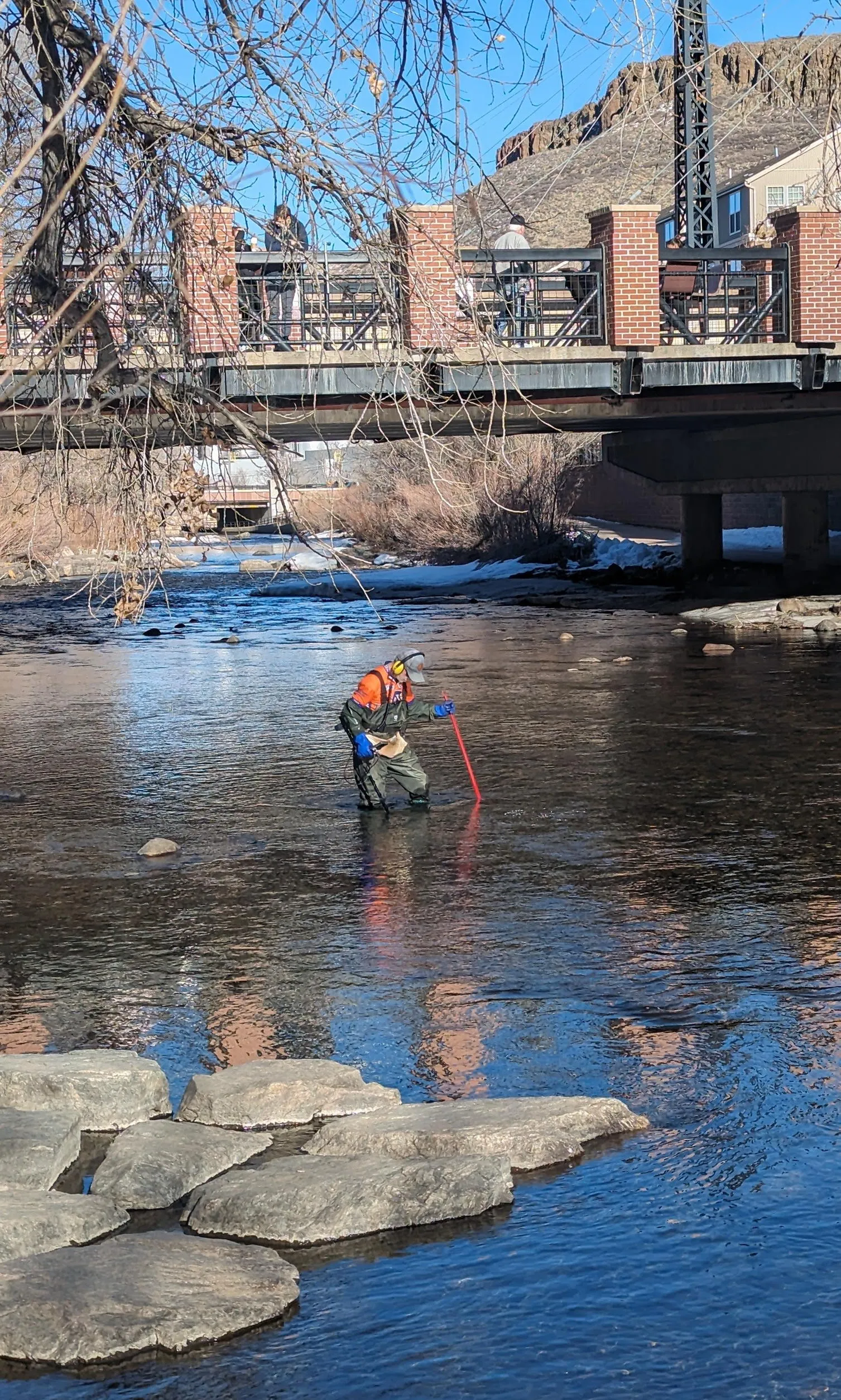 A man stands knee-deep in Clear Creek, using a metal detector to scan the bottom.