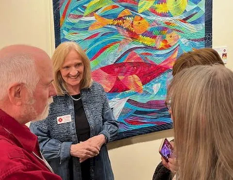Three women and one man in chatting in front of  a colorful quilt with fish motif.