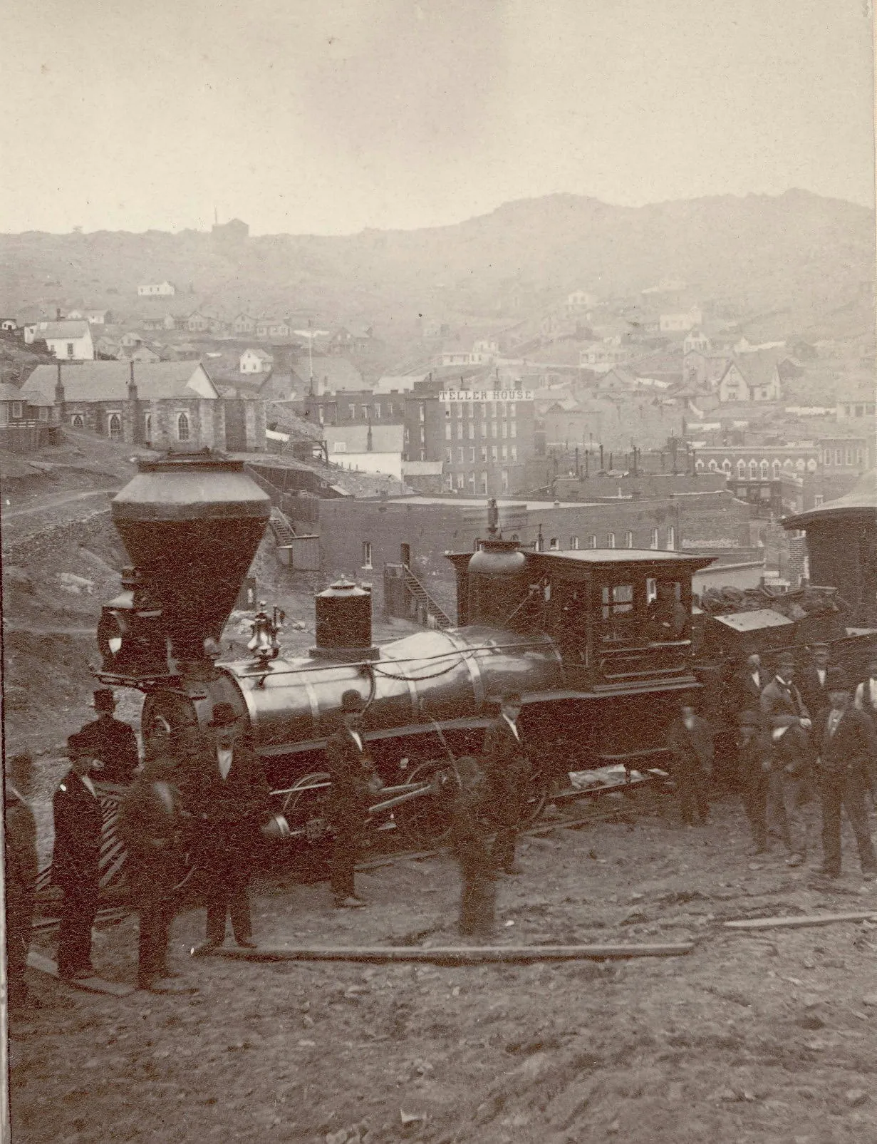 Men in suits and hats standing in front of a steam locomotive.  Mountainous Central City behind, including the Teller House.