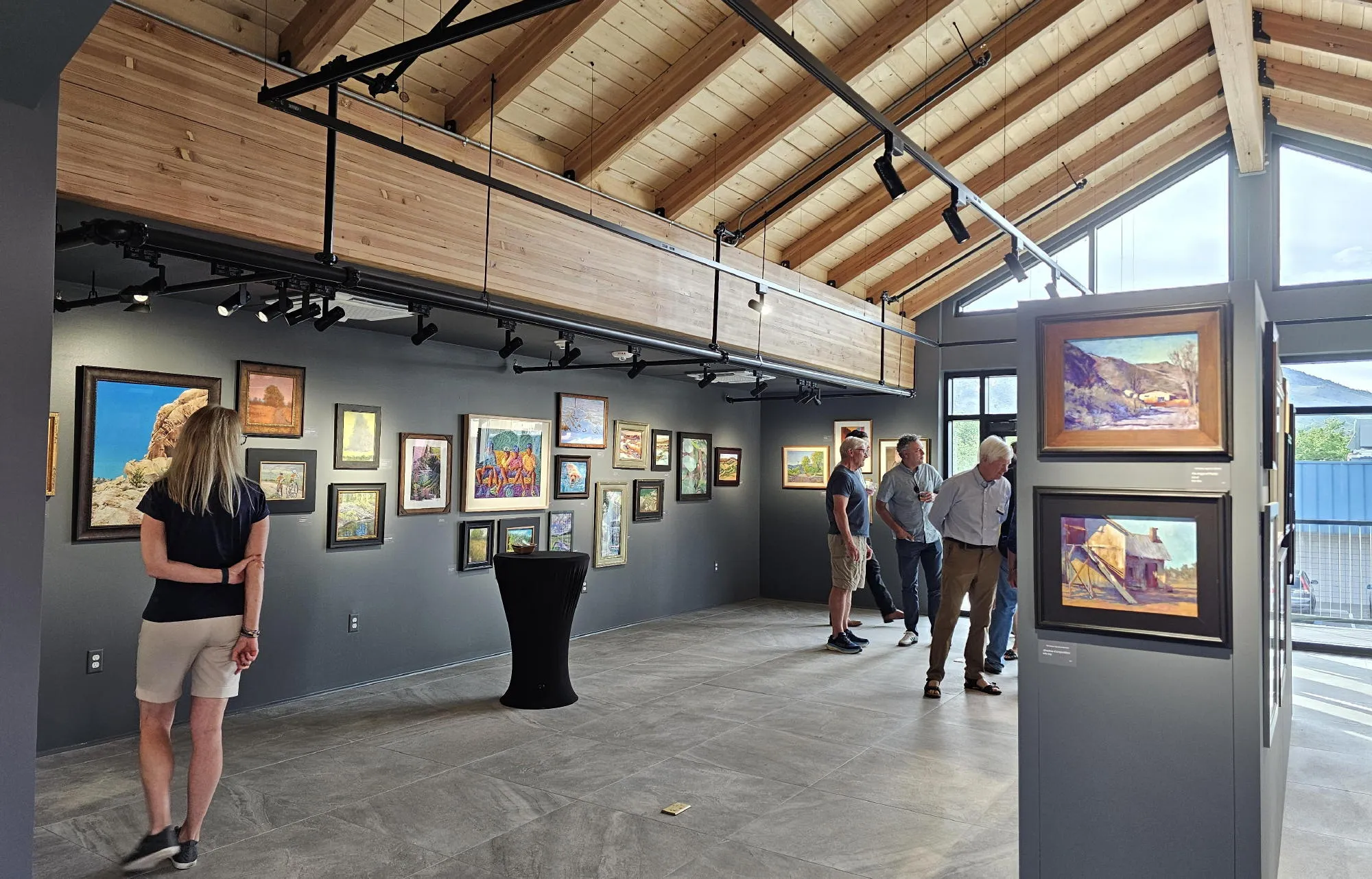 gallery room with vaulted timber ceiling, painting on the all, people, and large windows looking out at the foothills