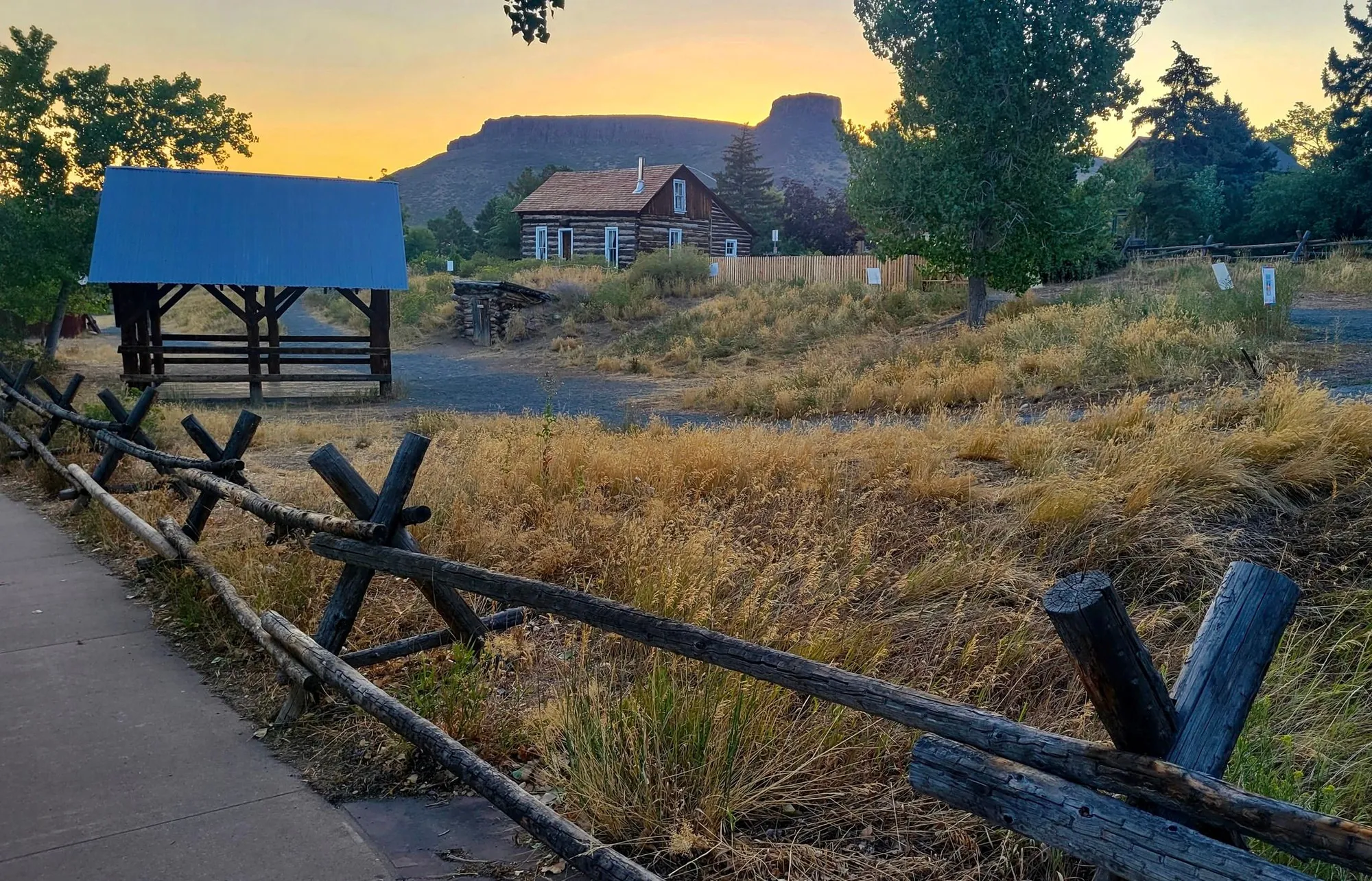 looking over the split rail fence at grass, a hay barn, a cabin, and castle rock in the background