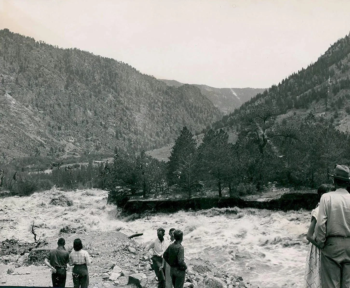 A group of people watch flood waters alongside a steep mountain slope - construction rubble along the bank.