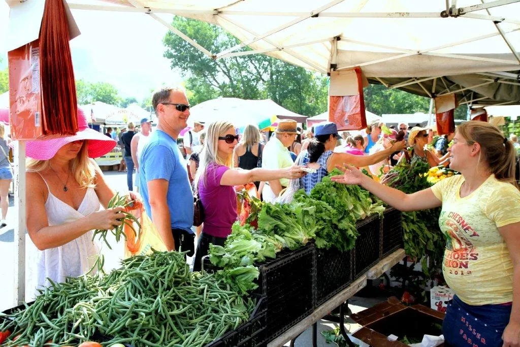 people lined up before a table of green vegetables, one loading a bag, one handing money across counter