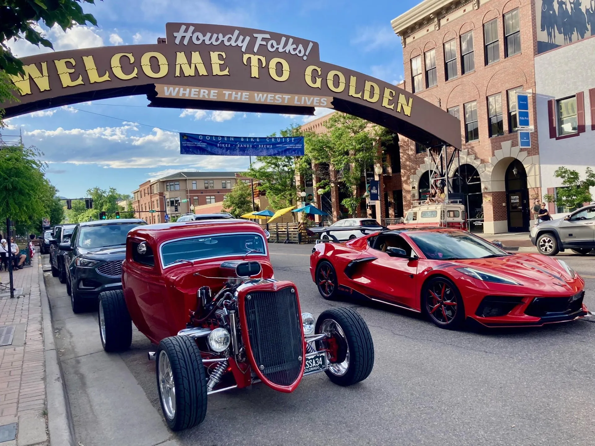 two red cars under the downtown welcome arch--a sports car in the driving lane and a Roadster parked on the side