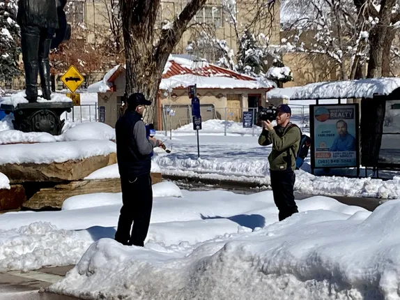 A reporter with microphone and a videographer stand in snow near Parfet Park