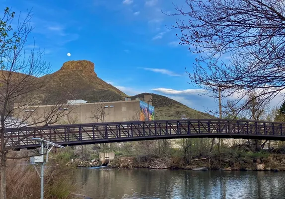View from Vanover Park - bridge in foreground, Coors and Castle Rock in background, moon overhead, budding trees on side