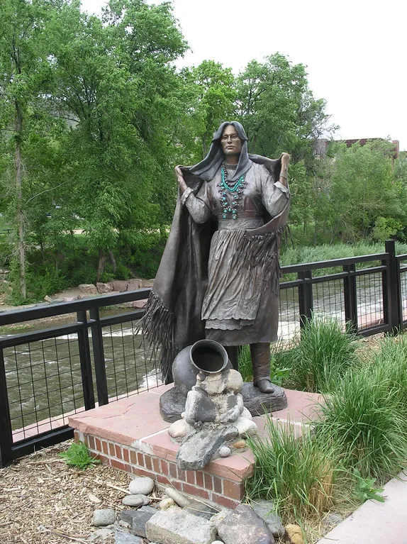 bronze statue of a native American woman wrapped in a fringed shawl or blanket, wearing turquoise necklaces