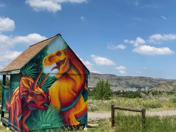 small wooden shed with a painting of a triceratops and a t-rex - South Table Mountain in background