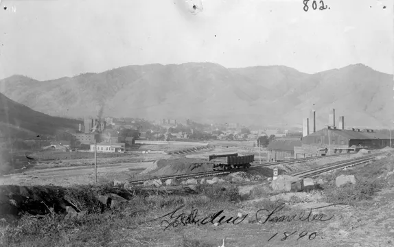 industrial-looking building with several smokestacks and railroad tracks with one flatbed car in the foreground