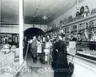 old-fashioned grocery store with merchandise behind the counter, two women patrons and five men, apparently employees